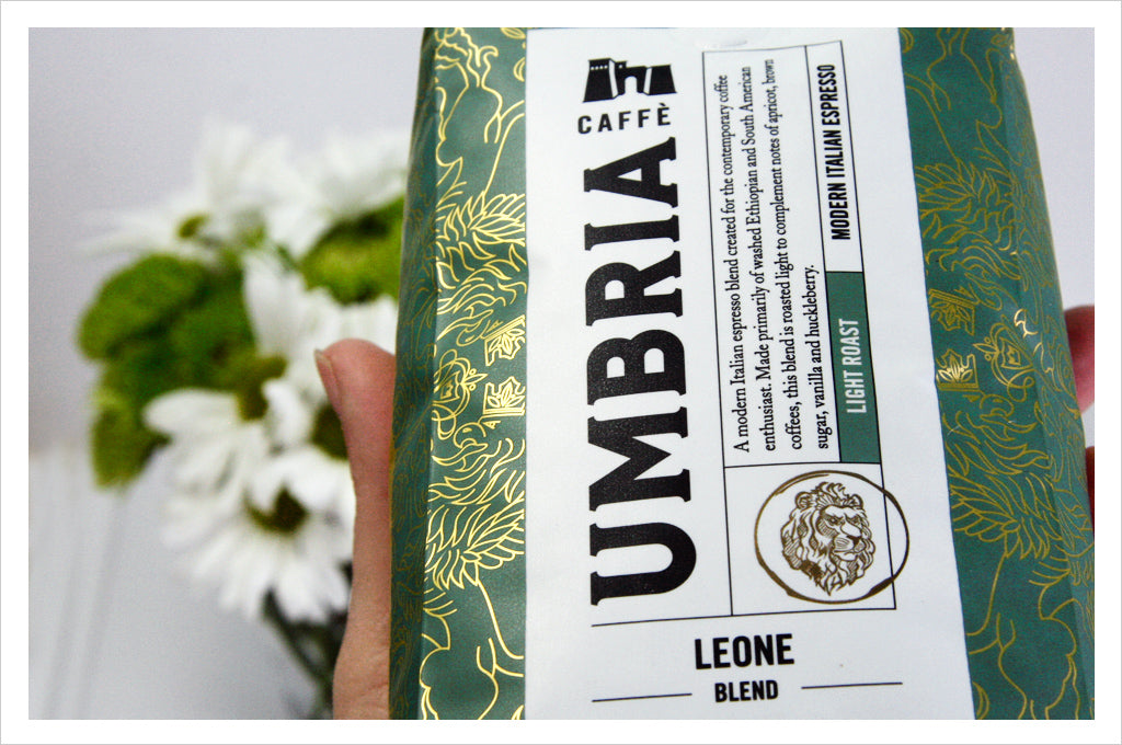 a hand holds a green bag of Leone blend coffee, green flowers in background