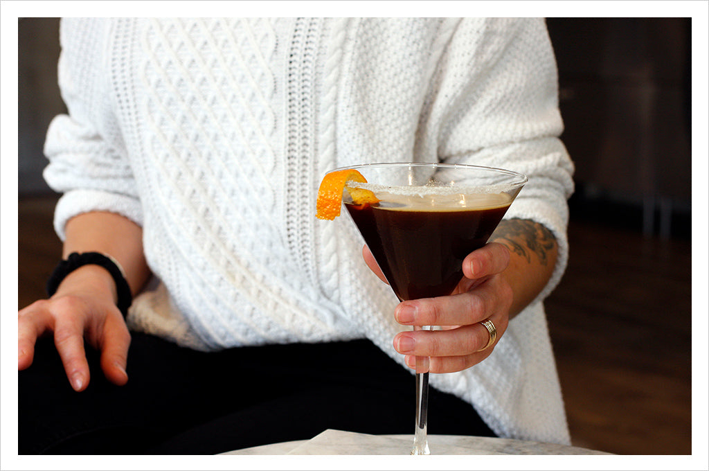 a woman in a white sweater holding a coffee martini with orange twist