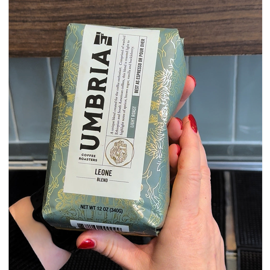 A hand with red painted nails holds a package of Umbria Coffee Roasters Leone Blend light roast coffee. The packaging is a sage green with gold floral patterns and a large white label on the front.