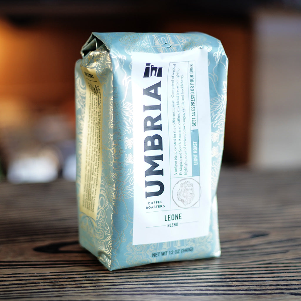 A bag of Umbria coffee beans, specifically the Leone Blend, standing on a dark wooden table. The packaging is a light green with ornate gold foil patterns and a large white label on the front.