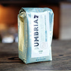 A bag of Umbria coffee beans, specifically the Leone Blend, standing on a dark wooden table. The packaging is a light green with ornate gold foil patterns and a large white label on the front.