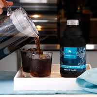coffee concentrate bottle and two glasses on tray, pitcher is pouring cold brew into glass